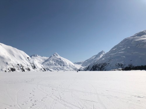 A vast snowy landscape with mountains under a clear blue sky, featuring a distant figure on the ice.