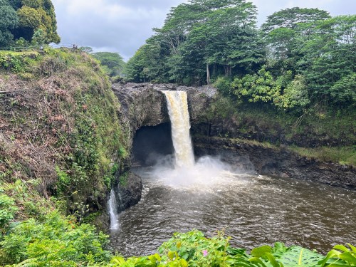 A waterfall cascades into a river surrounded by lush greenery and rocky cliffs under a cloudy sky.