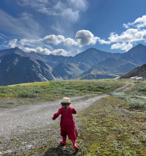 A child in a red dress and hat walks along a gravel path, surrounded by mountains and a blue sky with clouds.