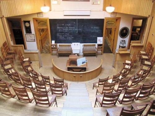A historic lecture hall with wooden benches, a central podium, and a chalkboard, set in a museum.