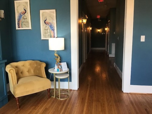 A cozy hallway with blue walls, a yellow armchair, a small table, and framed peacock art. Warm lighting enhances the space.