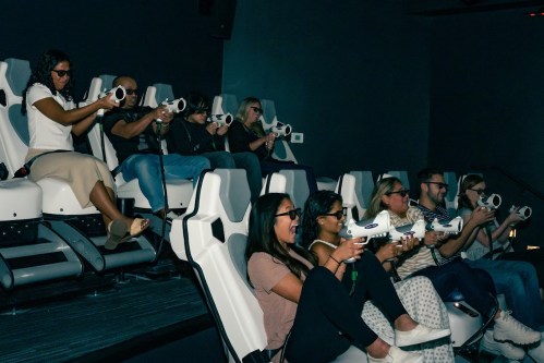 A group of people in 3D glasses seated in a theater, aiming toy guns at a screen during an interactive experience.