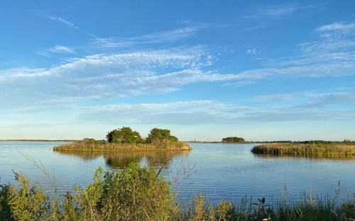 A serene landscape featuring calm water, small islands, and a clear blue sky with scattered clouds.