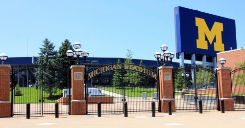 Entrance to Michigan Stadium, featuring a large "M" sign and landscaped grounds under a clear blue sky.