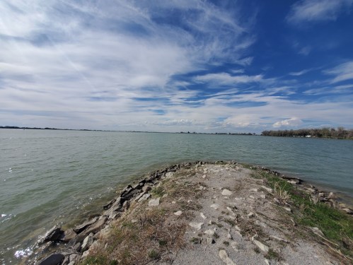 A rocky shoreline extends into a calm lake under a blue sky with scattered clouds.