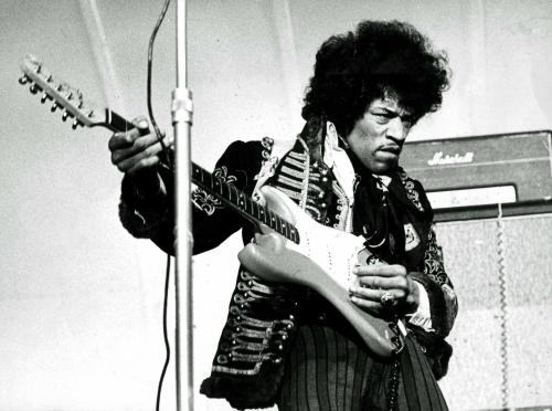 A musician in a flamboyant outfit plays an electric guitar on stage, with amplifiers in the background.