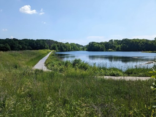 A serene lake surrounded by lush greenery and a winding path under a clear blue sky.
