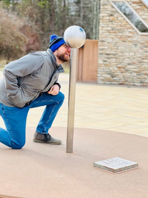 A person kneels beside a metal sculpture, peering into a spherical opening at the top.