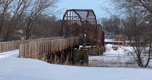 A rusty, old bridge spans a snowy landscape, with trees in the background and a wooden walkway leading to it.