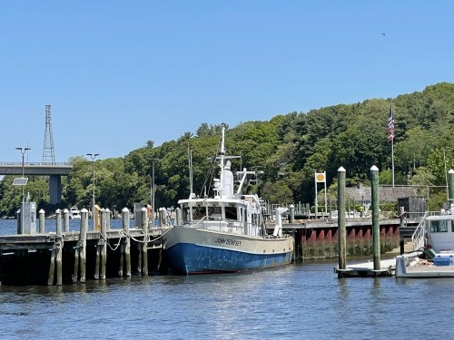 A boat named "John Victory" docked at a marina with green trees and a bridge in the background under a clear blue sky.