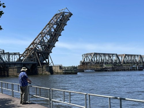 A man stands by the water, observing a raised drawbridge against a clear blue sky.