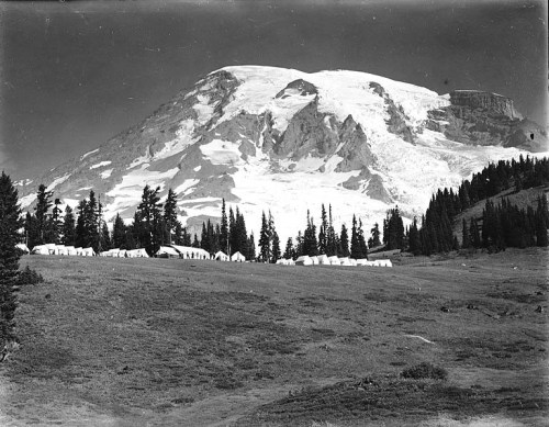 A black and white photo of a mountain with snow-capped peaks, surrounded by trees and a row of tents in the foreground.