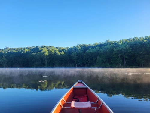 A canoe on a calm lake with mist rising, surrounded by lush green trees under a clear blue sky.