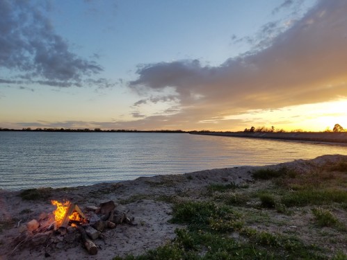 A serene lakeside scene at sunset, featuring a small campfire on the sandy shore and calm water reflecting the sky.
