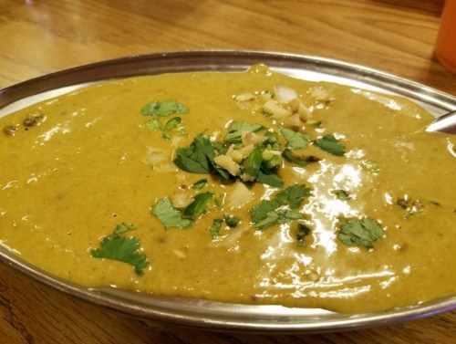A silver platter holds a vibrant green dish topped with chopped herbs and nuts, served on a wooden table.