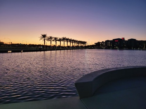 A serene lake at sunset, with palm trees lining the shore and buildings in the background against a colorful sky.