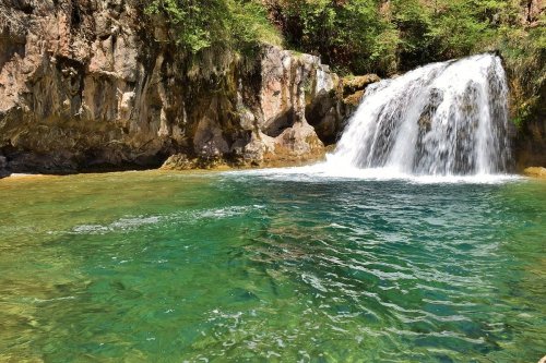 A serene waterfall cascades into a clear, turquoise pool surrounded by lush greenery and rocky cliffs.