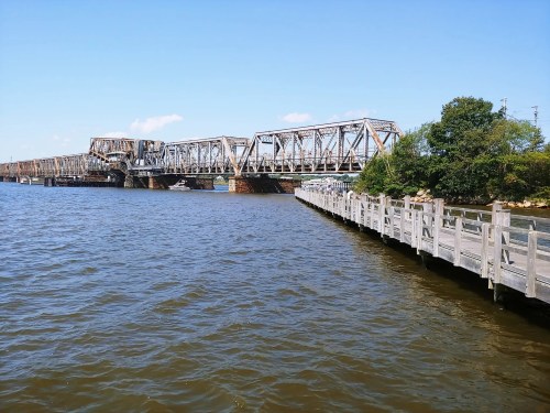 A metal bridge spans a river, with a wooden pier extending along the water's edge under a clear blue sky.