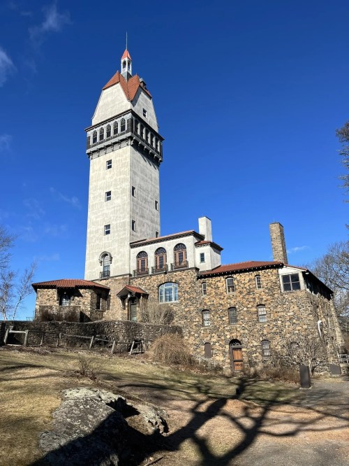 Historic stone building with a tall tower against a clear blue sky, surrounded by bare trees and grassy grounds.