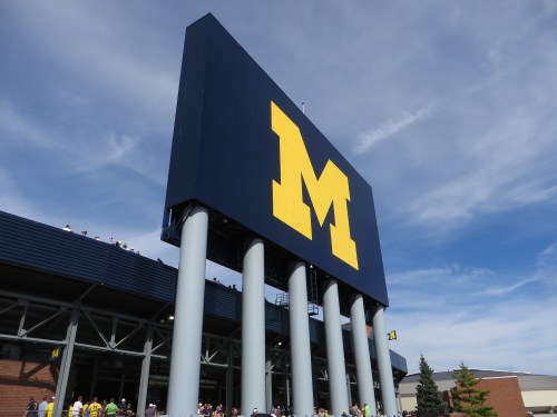 Large blue sign with a yellow "M" at a stadium, under a clear blue sky with spectators in the background.