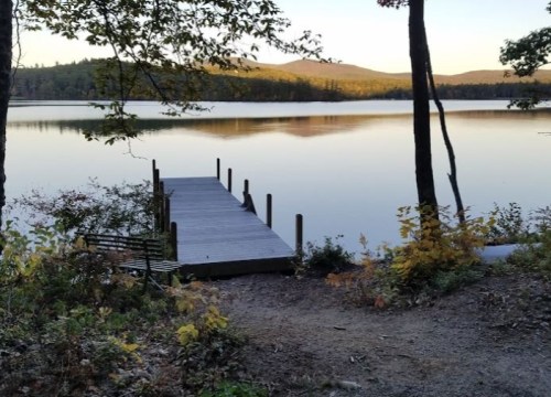 A serene lake view with a wooden dock, surrounded by trees and mountains in the background during sunset.