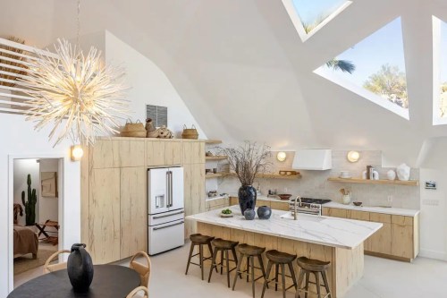 Modern kitchen with wood cabinetry, marble island, and unique light fixture, featuring large windows and a bright atmosphere.