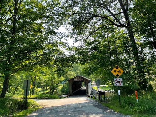 A covered bridge surrounded by lush green trees, with road signs indicating height limit and no entry.