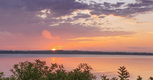 A serene sunset over a calm lake, with colorful clouds reflecting on the water and greenery in the foreground.