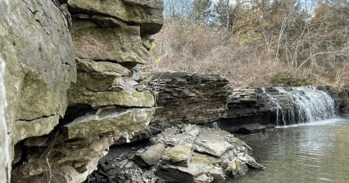 A rocky cliffside beside a calm water pool, with a small waterfall cascading into the water, surrounded by trees.