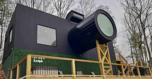 A modern black house with a cylindrical window, surrounded by trees and featuring a wooden deck.