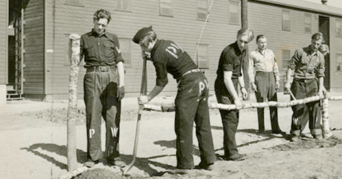 Historical photo of men working together to dig a trench, with buildings in the background.