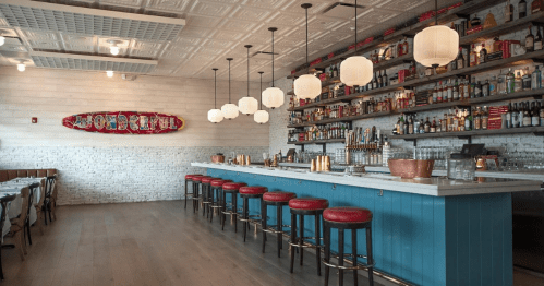 A stylish restaurant interior featuring a bar with stools, shelves of drinks, and decorative lighting.