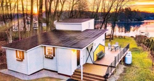 A cozy lakeside house at sunset, surrounded by trees, with a deck and outdoor seating.