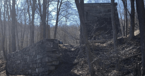 A stone structure partially hidden by trees, with a larger stone building in the background on a hillside.