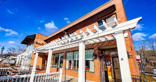 A brick restaurant with a white awning, surrounded by outdoor seating, under a blue sky with scattered clouds.