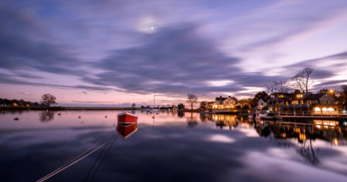 A serene lake at dusk with a red boat and reflections of clouds and houses on the water's surface.