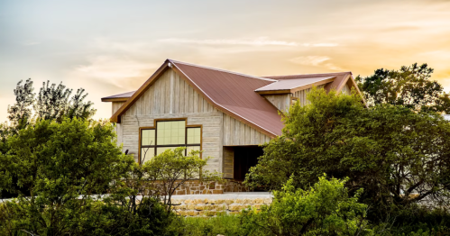 A modern wooden house with a metal roof, surrounded by greenery, under a colorful sunset sky.