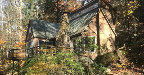 A cozy cabin nestled in a forest, surrounded by autumn foliage and greenery.