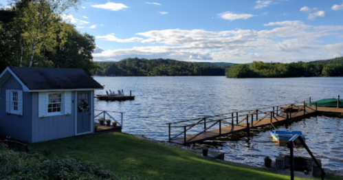 A serene lakeside view featuring a small blue shed, a wooden dock, and lush green surroundings under a clear sky.