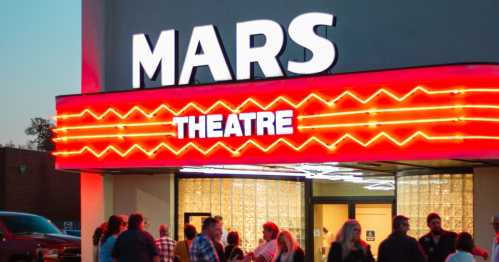 A crowd gathers outside the Mars Theatre, featuring a bright neon sign and a vibrant atmosphere at dusk.