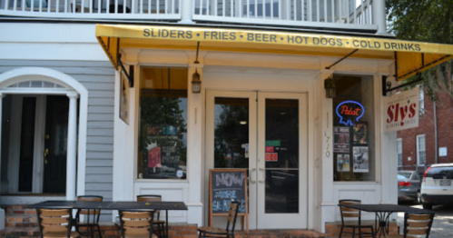 Exterior of a casual eatery with a yellow awning, outdoor seating, and a menu featuring sliders, fries, and drinks.