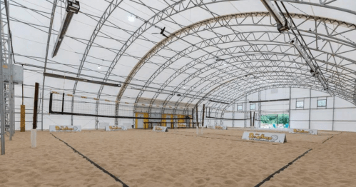 Indoor sand volleyball court with a large, white tent structure and multiple nets set up on a sandy floor.
