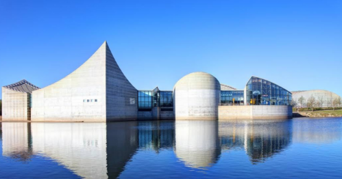 Modern architectural building with unique shapes, reflecting in calm water under a clear blue sky.
