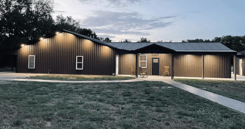 A modern brown metal building with a porch, surrounded by grass and a pathway, under a twilight sky.