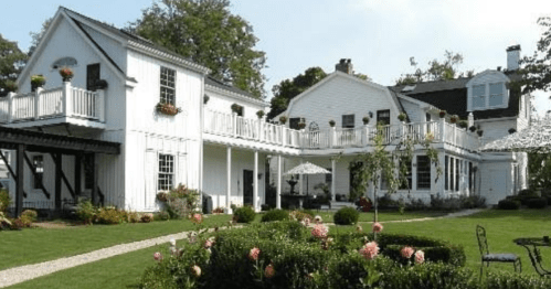 A charming white house with balconies, surrounded by a lush garden and patio furniture on a sunny day.