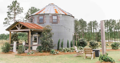 A rustic, round metal building with a sloped roof, surrounded by greenery and wooden seating in a serene outdoor setting.