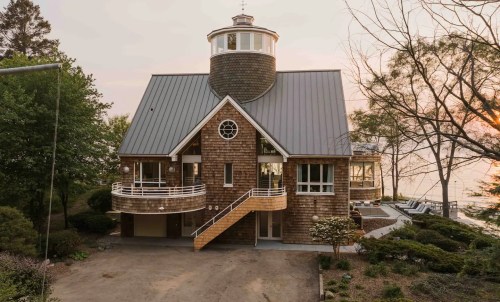 A unique waterfront house with a lighthouse-style tower, surrounded by trees and a serene landscape at sunset.