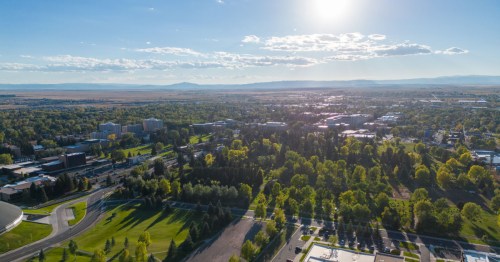 Aerial view of a green campus with buildings, trees, and mountains in the background under a bright blue sky.