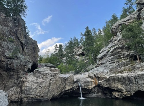 A serene landscape featuring a waterfall cascading into a rocky pool, surrounded by trees and cliffs under a blue sky.