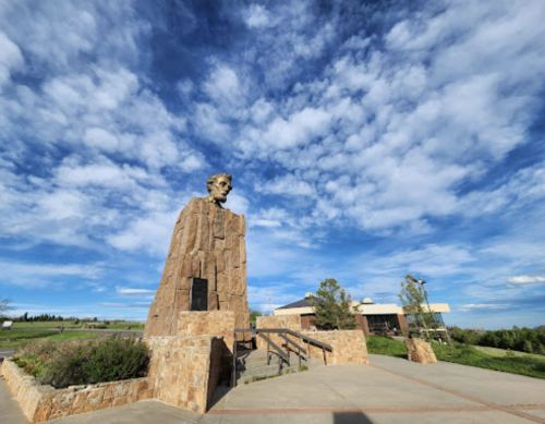 A large stone monument with a bust atop, set against a blue sky with clouds, surrounded by greenery.